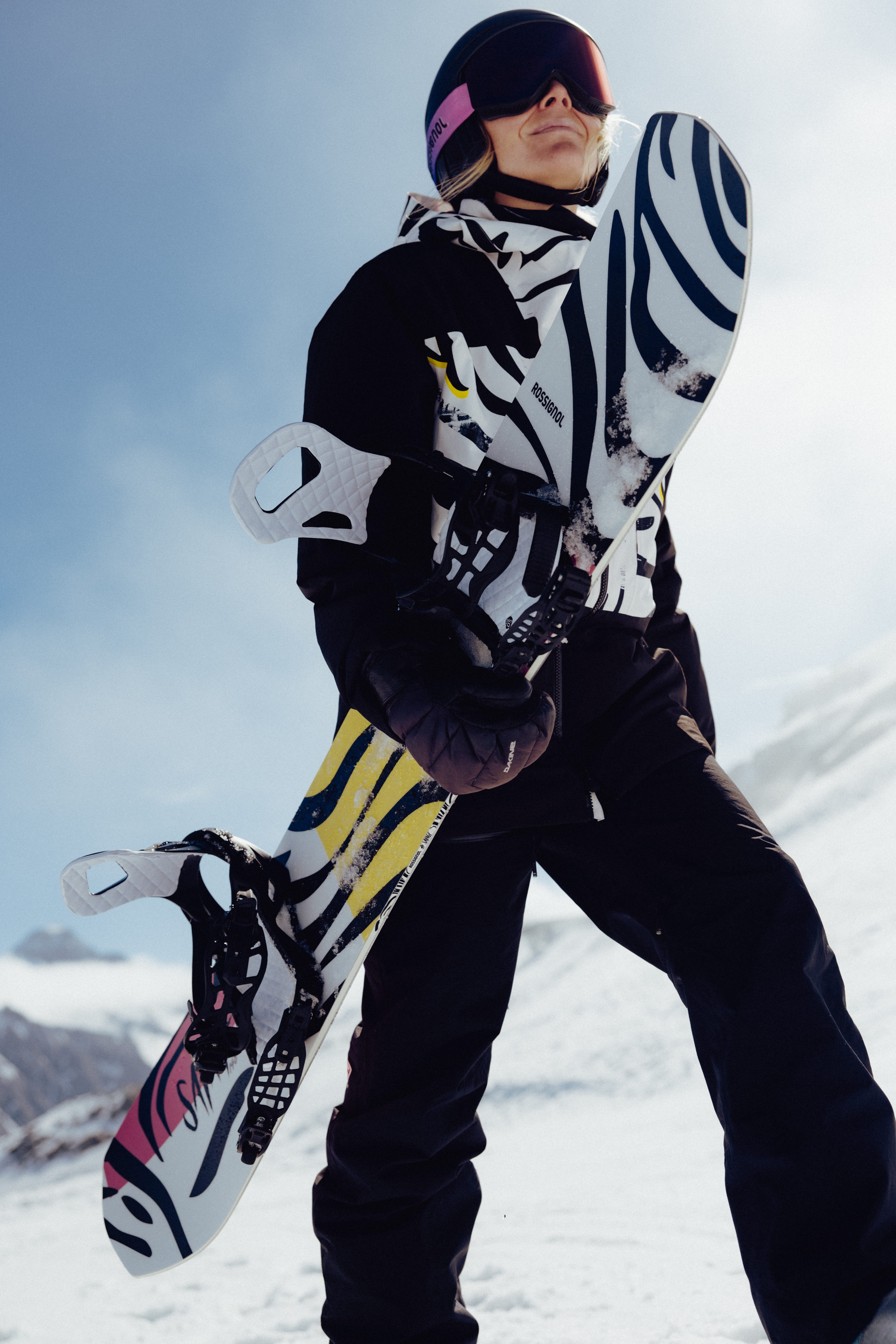 Personne debout sur une pente enneigée en montagne, tenant un snowboard aux graphismes noirs et blancs audacieux, portant une tenue de neige, un casque et un masque, avec un paysage montagneux en arrière-plan.