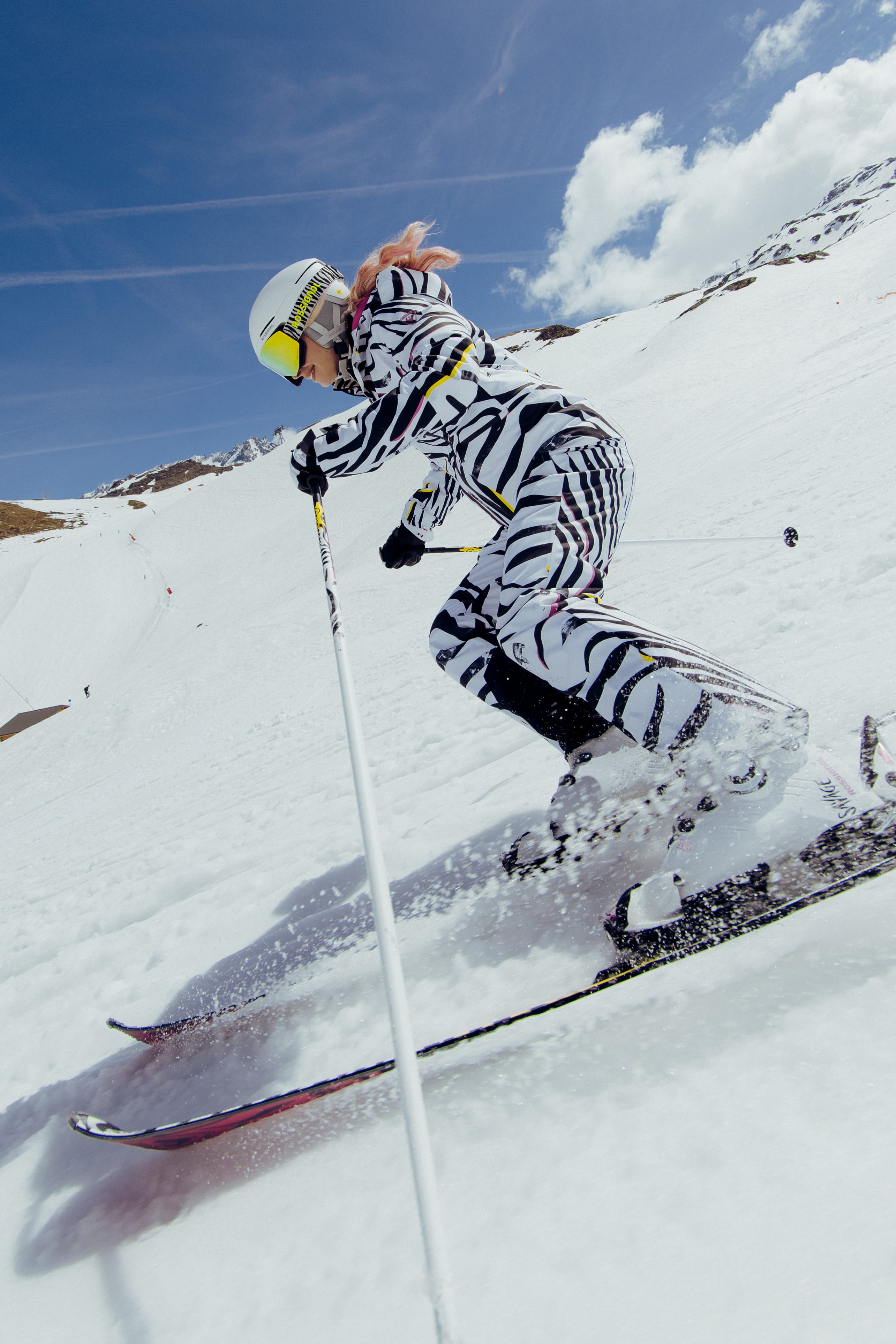 Action shot of a skier in a bold black-and-white patterned ski outfit skiing through a snowy slope, with snow spraying from the skis.