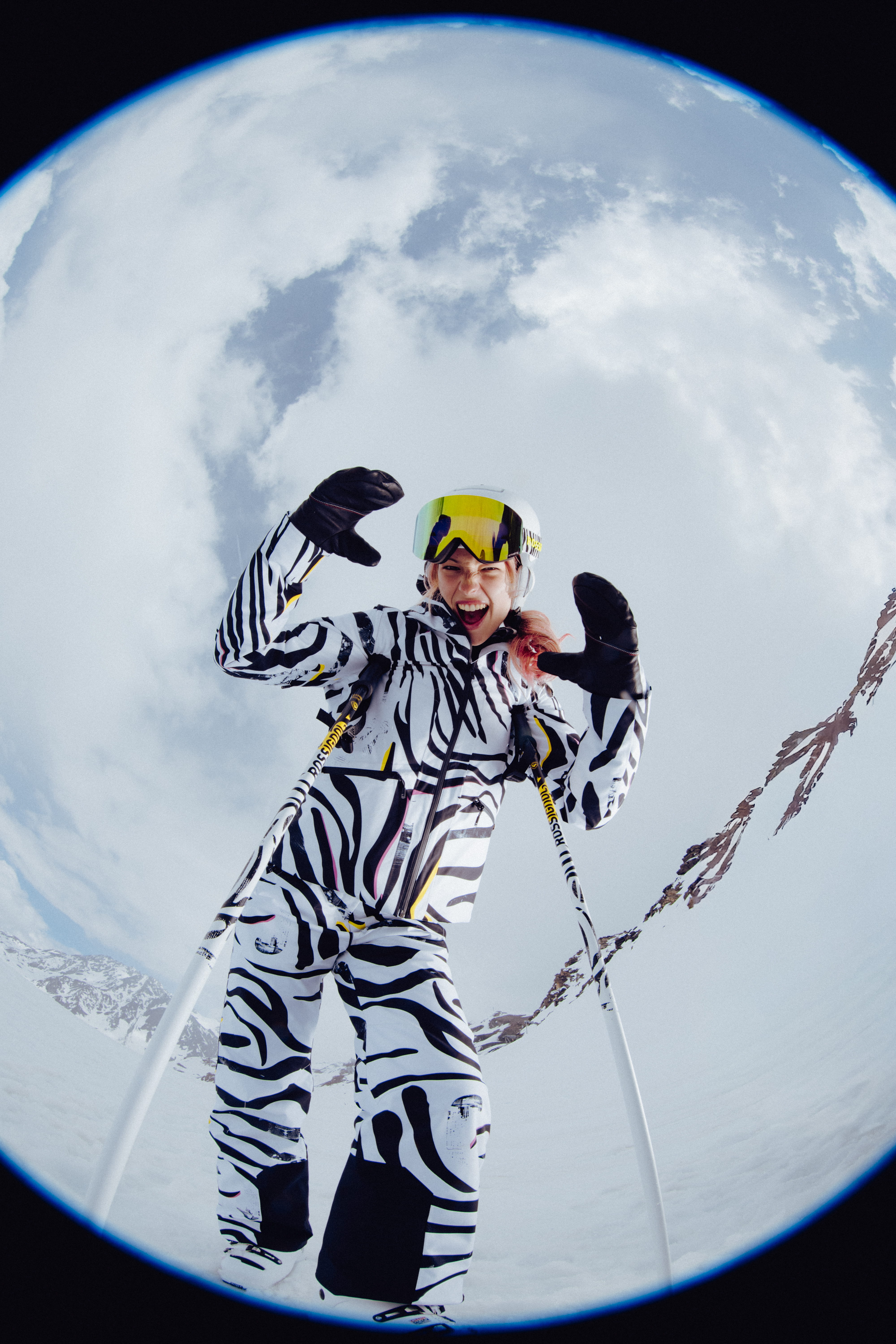 Fisheye view of a skier in a bold black-and-white patterned ski outfit standing on a snowy mountain slope with ski poles, wearing a white helmet and mirrored goggles beneath a cloudy sky.