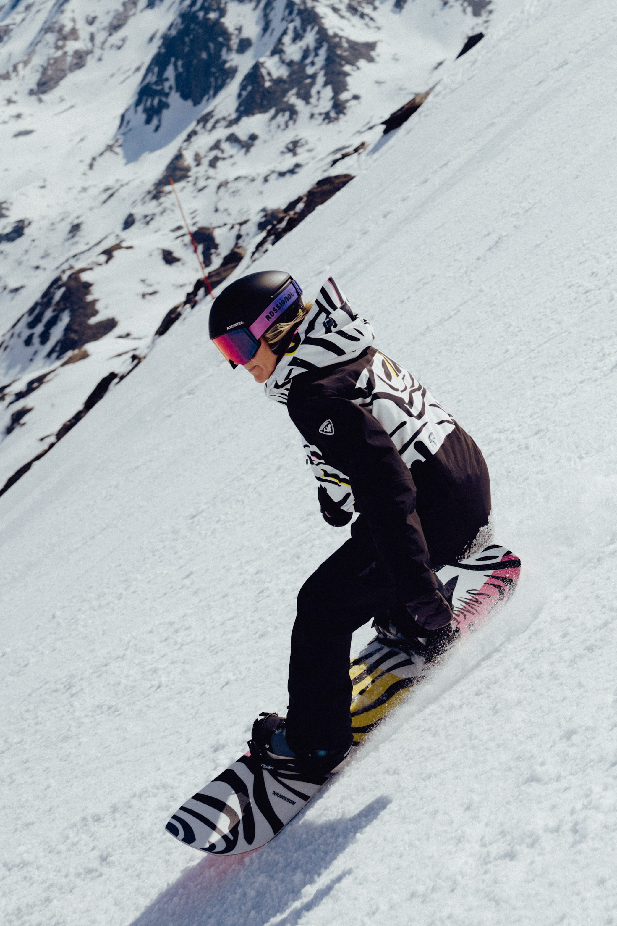 Snowboardeur descendant une pente enneigée, vêtu d’une tenue noire et blanche à motif graphique et d’un masque à écran miroir.